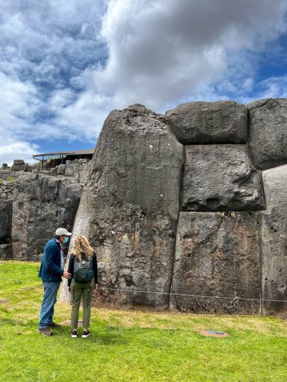 Two people, one wearing a backpack, stand next to a large ancient stone wall under a partly cloudy sky, discovering the timeless beauty of Peru's Sacred Valley.