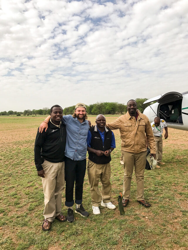 Four people standing and smiling, with arms around each other's shoulders, outside next to a small airplane on a grassy field under a cloudy sky, ready for their Wildlife Safari in Tanzania.