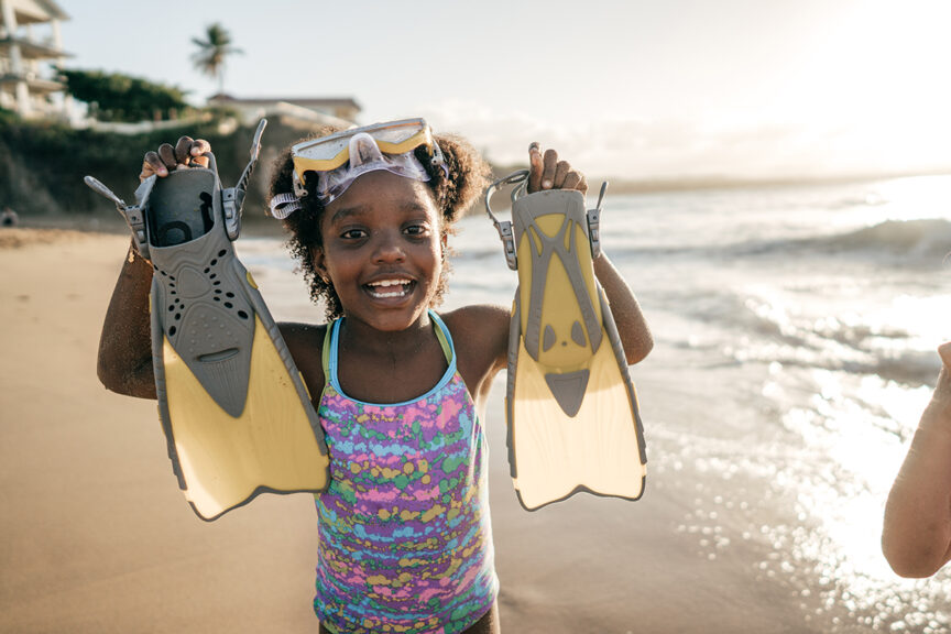 A child stands on a beach, smiling and holding a pair of snorkeling fins up to the camera. The sun is setting in the background, and waves lap at the shore, making it a perfect scene for any snorkeling trip. Don’t forget to add fins to your list of things to pack!