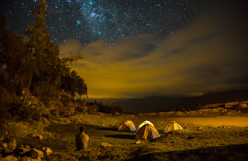 A person sits near three illuminated tents under a starry night sky in the wilds of Peru, surrounded by trees and rocky terrain, celebrating their 60th birthday.