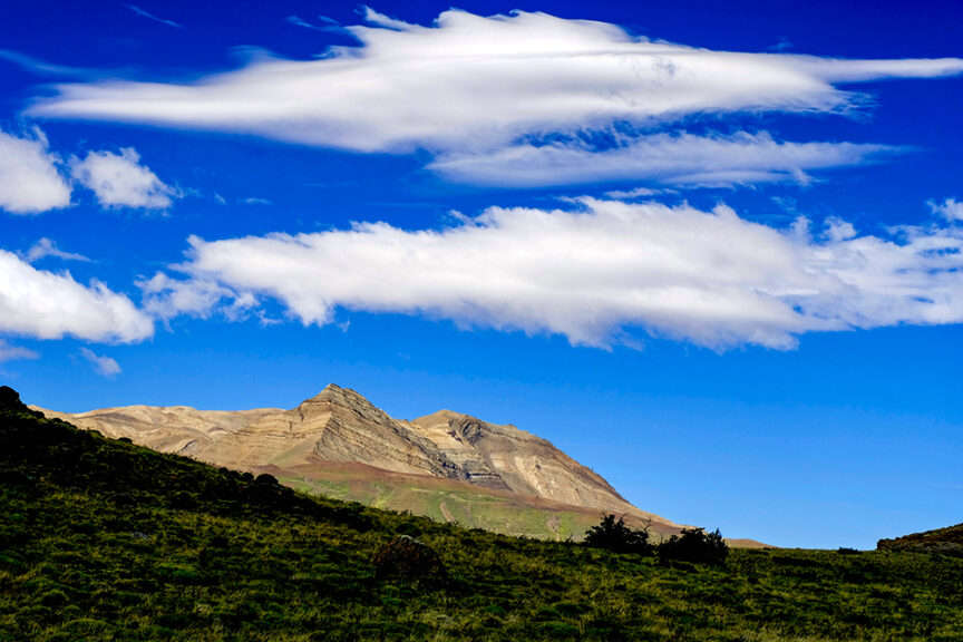 A stunning impression of Patagonia unfolds before you: a mountain range under a clear blue sky with white clouds, foregrounded by a verdant green landscape.