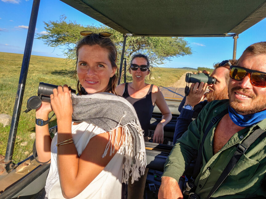 Four people are on a Serengeti Safari vehicle, looking through binoculars and cameras, surrounded by a grassy landscape under a clear blue sky.