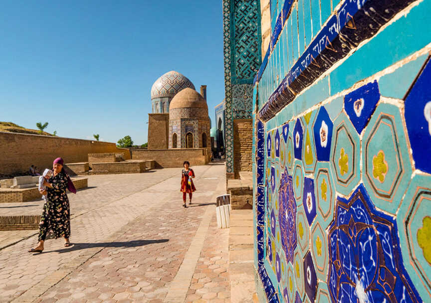 Two people walking on a sunlit path near ornate, blue-tiled structures with dome roofs in a historical site, embarking on a cultural journey along the ancient Silk Road.