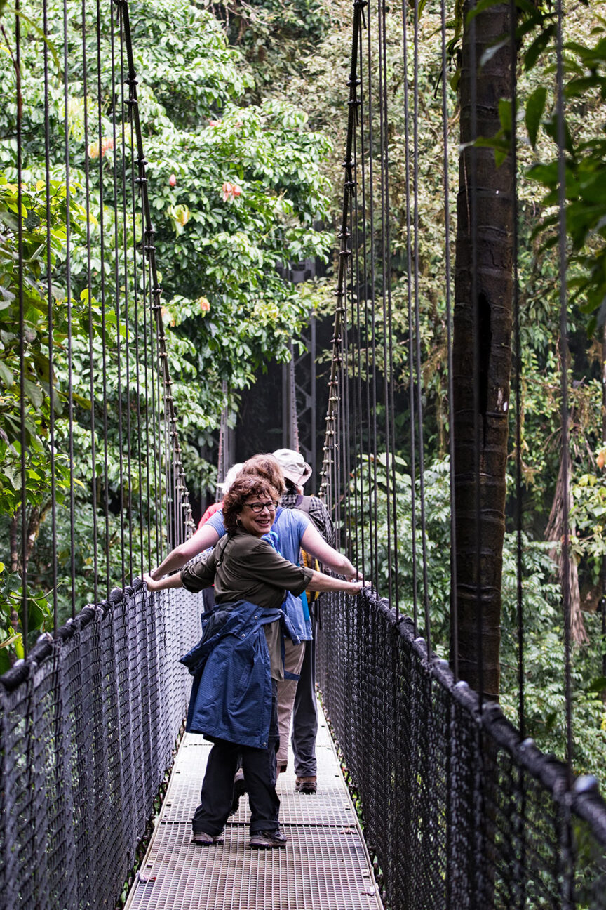 Three individuals crossing a narrow suspension bridge surrounded by lush green trees in Costa Rica. The person in the foreground smiles and turns back, while the others face forward. The scene is teeming with life, almost as if colorful creatures could pop out from the foliage any moment.