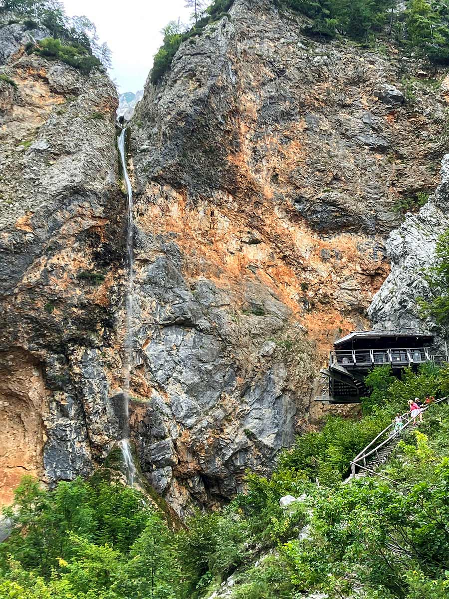 A small waterfall cascades down a rocky cliff near a wooden structure with stairs leading to it, surrounded by lush greenery. Visit this serene spot in Slovenia for one of the most beautiful places you'll ever experience.