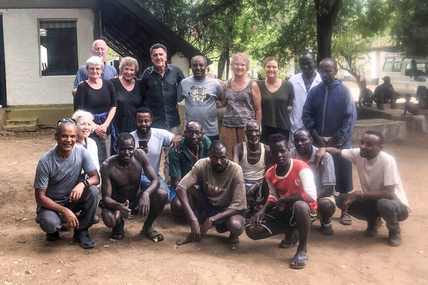 A group of people, some standing and some kneeling, pose for a photo outdoors in front of a building. Trees and other individuals are visible in the background. They are part of the Wilderness Travelers, capturing memories despite challenges posed by the pandemic.