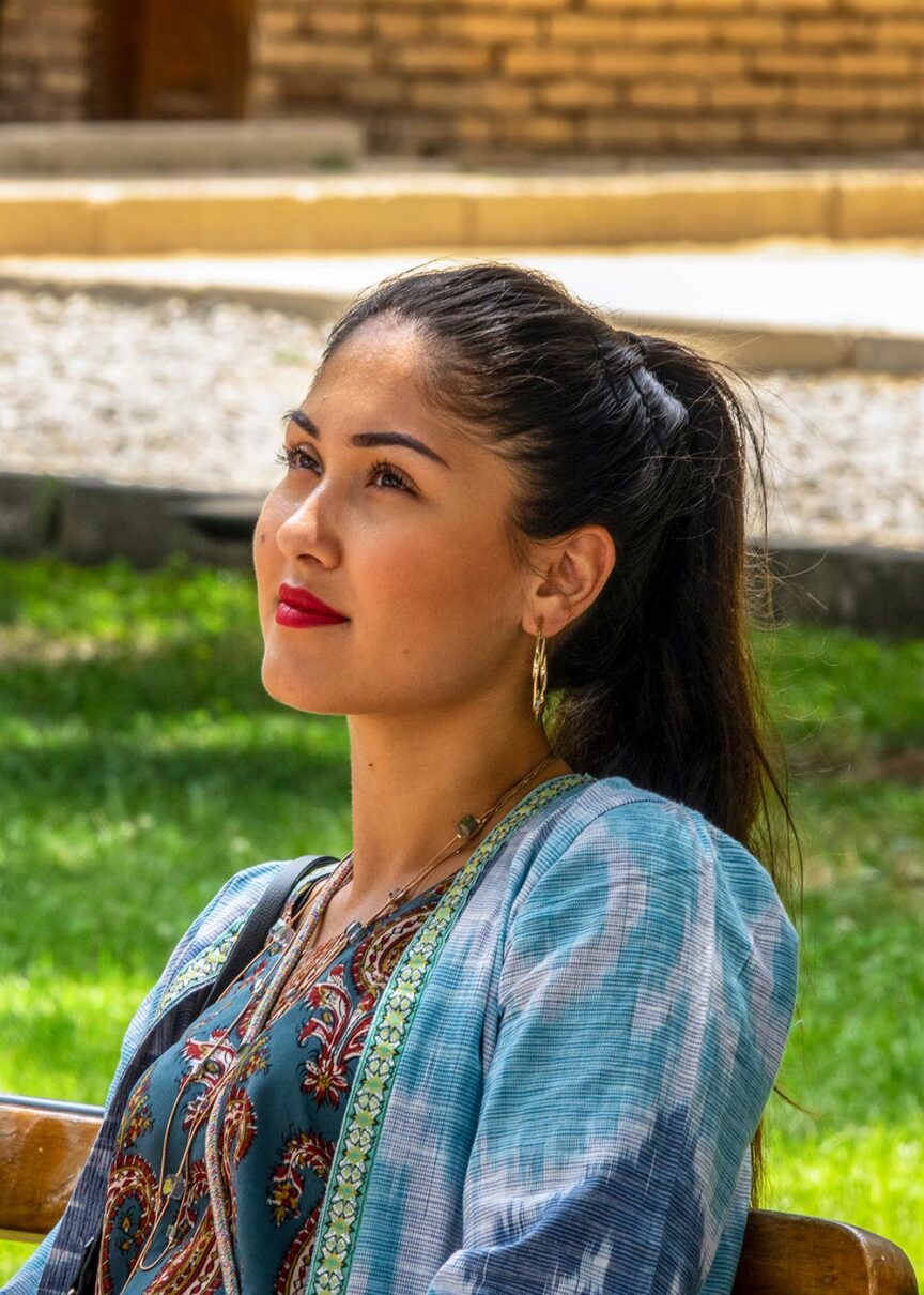 A woman with a ponytail and red lipstick sits on a bench, looking upward. She is wearing a blue patterned top with embroidered detailing, reminiscent of designs found along the Silk Road. The background shows grass, a stone walkway, and a brick structure, adding to the sense of a cultural journey.