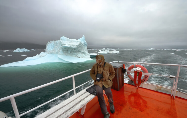 A person in outdoor clothing sits on a boat's railing, with large icebergs floating in the surrounding cold water under cloudy Arctic skies.
