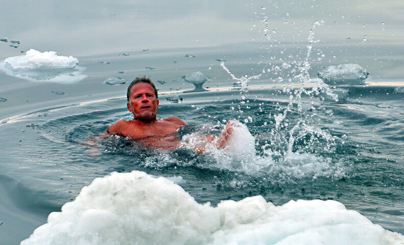 A man swims in the icy waters of the Arctic, surrounded by floating ice chunks and snow, creating splashes as he moves.