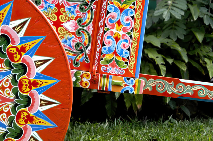 A vividly painted wooden cart featuring intricate floral and geometric designs, reminiscent of the whimsical style in "Where the Wild Things Are," is situated outdoors against a backdrop of lush green foliage.