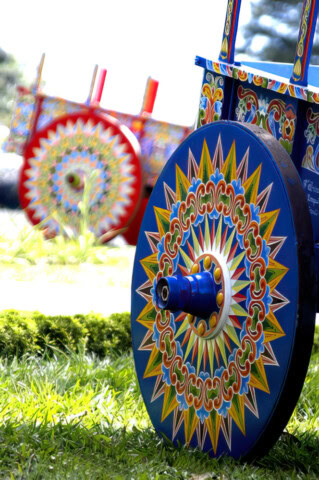 Close-up of intricately painted wooden cart wheels in vibrant colors with geometric and floral patterns, reminiscent of "Where the Wild Things Are," displayed outdoors on grass.