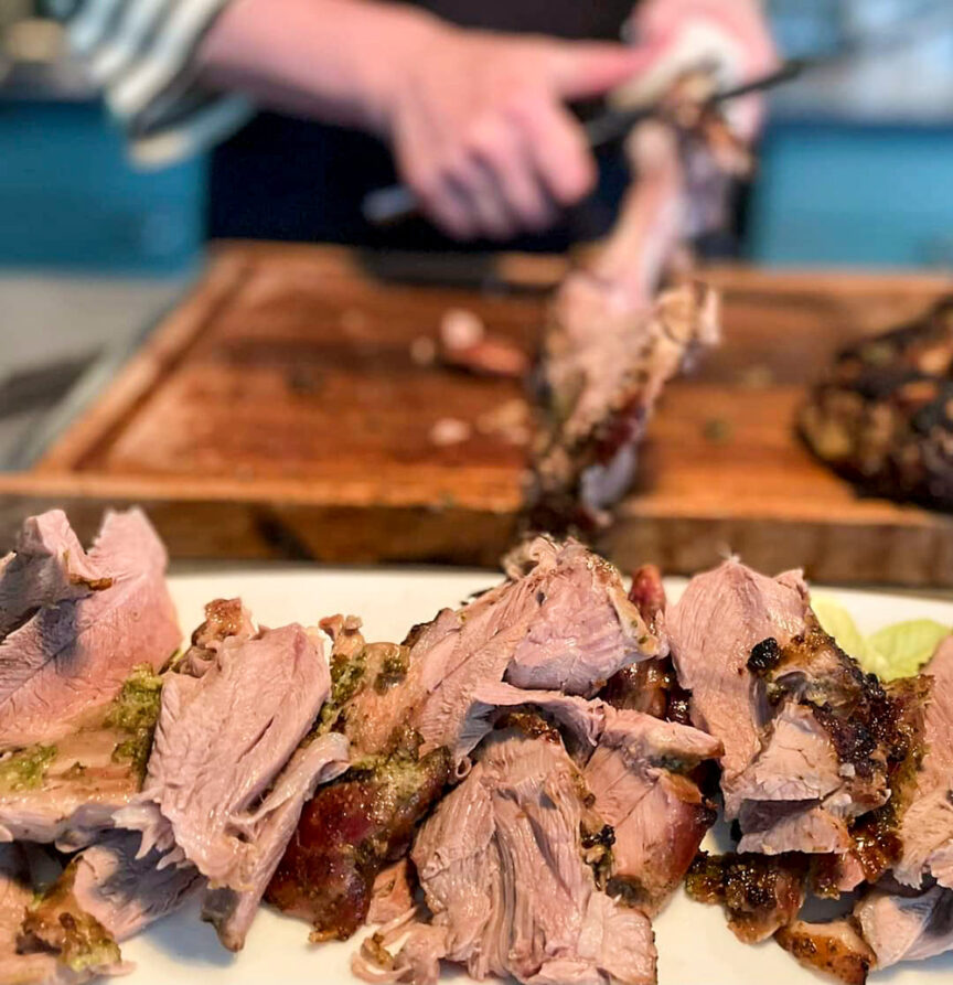 A person slices roasted meat on a wooden cutting board in the background, with a plate of carved meat pieces reflecting the essence of a Faroe Islands feast in the foreground.