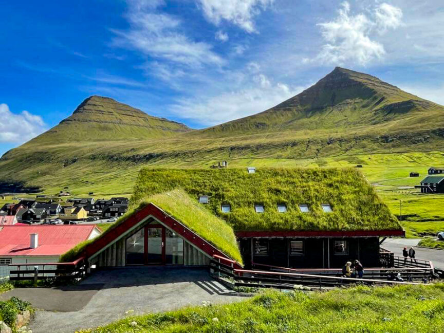 A building with a grass-covered roof stands against a backdrop of green hills and distant mountain peaks under a blue sky with scattered clouds. The scene evokes the serene beauty of the Faroe Islands, with reflections of nearby houses visible in the clear water.