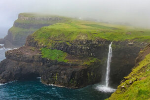 A cliffside waterfall cascades into the ocean under foggy skies in the Faroe Islands. The landscape is green and rugged, with a few buildings visible in the background, perfect for travel reflections.