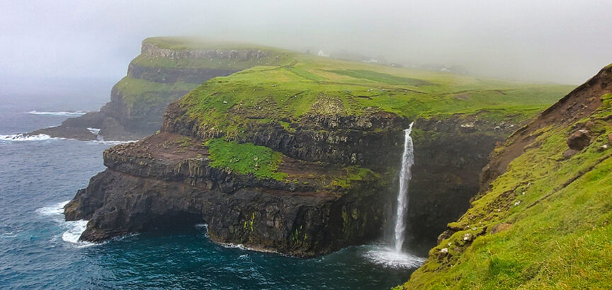 A cliffside waterfall cascades into the ocean under foggy skies in the Faroe Islands. The landscape is green and rugged, with a few buildings visible in the background, perfect for travel reflections.