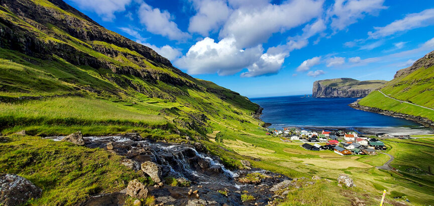 A small coastal village with colorful houses is nestled between green hills and cliffs, overlooking a calm blue sea under a partly cloudy sky, reminiscent of the serene landscapes found in the Faroe Islands.