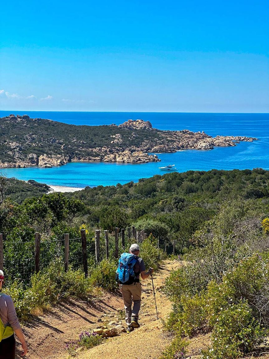 A person wearing a backpack hikes along a trail overlooking the scenic coastal landscape of Sardinia, with its clear blue waters, rocky outcrops, and lush greenery under a bright blue sky.