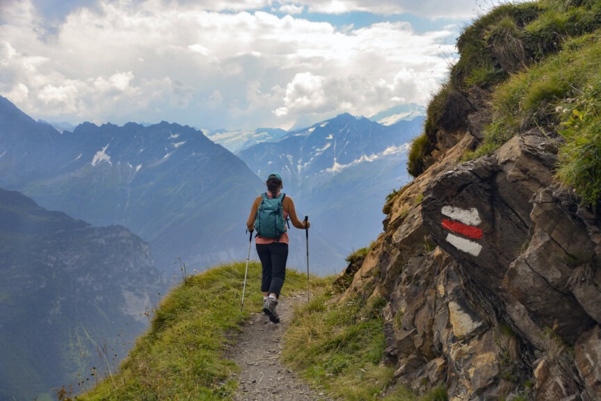 A hiker journeys along a narrow mountain trail in the Alps with trekking poles and a backpack. A red and white trail marker is visible on a rock, guiding the way. Snow-capped peaks loom majestically in the background, inviting an unforgettable hiking adventure.