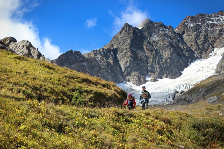 Two hikers with backpacks walk on a grassy slope adorned with yellow flowers in the majestic Alps, against a backdrop of rocky mountains and a glacier beneath a clear blue sky.
