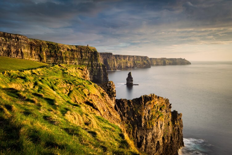 Sunlit Cliffs of Moher in Ireland, with lush green grass overlooking a calm sea, set against a partly cloudy sky—a true gem of the best region to visit.