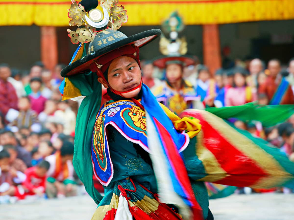 At a vibrant outdoor event, a person in traditional colorful attire performs a cultural dance reminiscent of Bhutan's Tshechu Festivals, captivating the crowd gathered to watch this spectacular celebration.