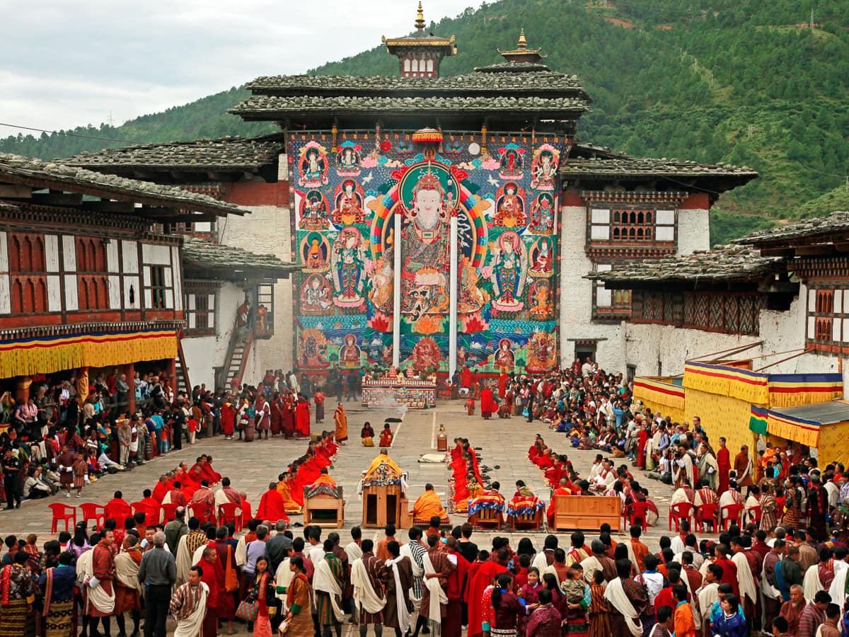 A large gathering of people at the vibrant Tshechu Festival in Bhutan, featuring monks and a colorful tapestry in a courtyard surrounded by traditional architecture and mountains.