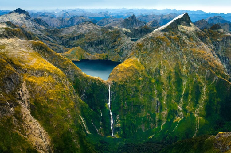 Aerial view of a mountainous landscape with a central lake, surrounded by rugged peaks. A waterfall flows from the lake, descending through lush greenery. This breathtaking scene captures the essence of New Zealand, a must-visit destination on any traveler's list.