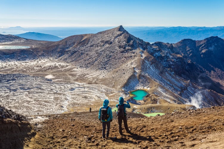 Two hikers stand on a rocky terrain, overlooking a mountainous landscape reminiscent of New Zealand's best islands, where snow patches and small turquoise lakes lie under a clear blue sky, inviting you to visit this natural wonder.