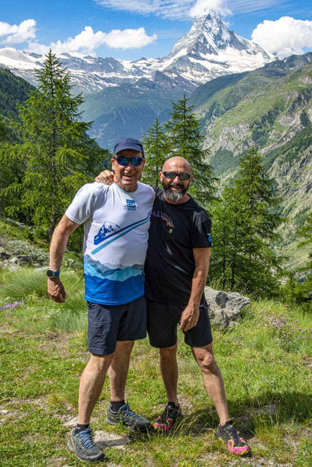 Two men smiling with arms around each other, standing in a mountainous area near Monte Rosa with the Matterhorn in the background, surrounded by green trees and grass, perhaps after a rewarding day of hiking.