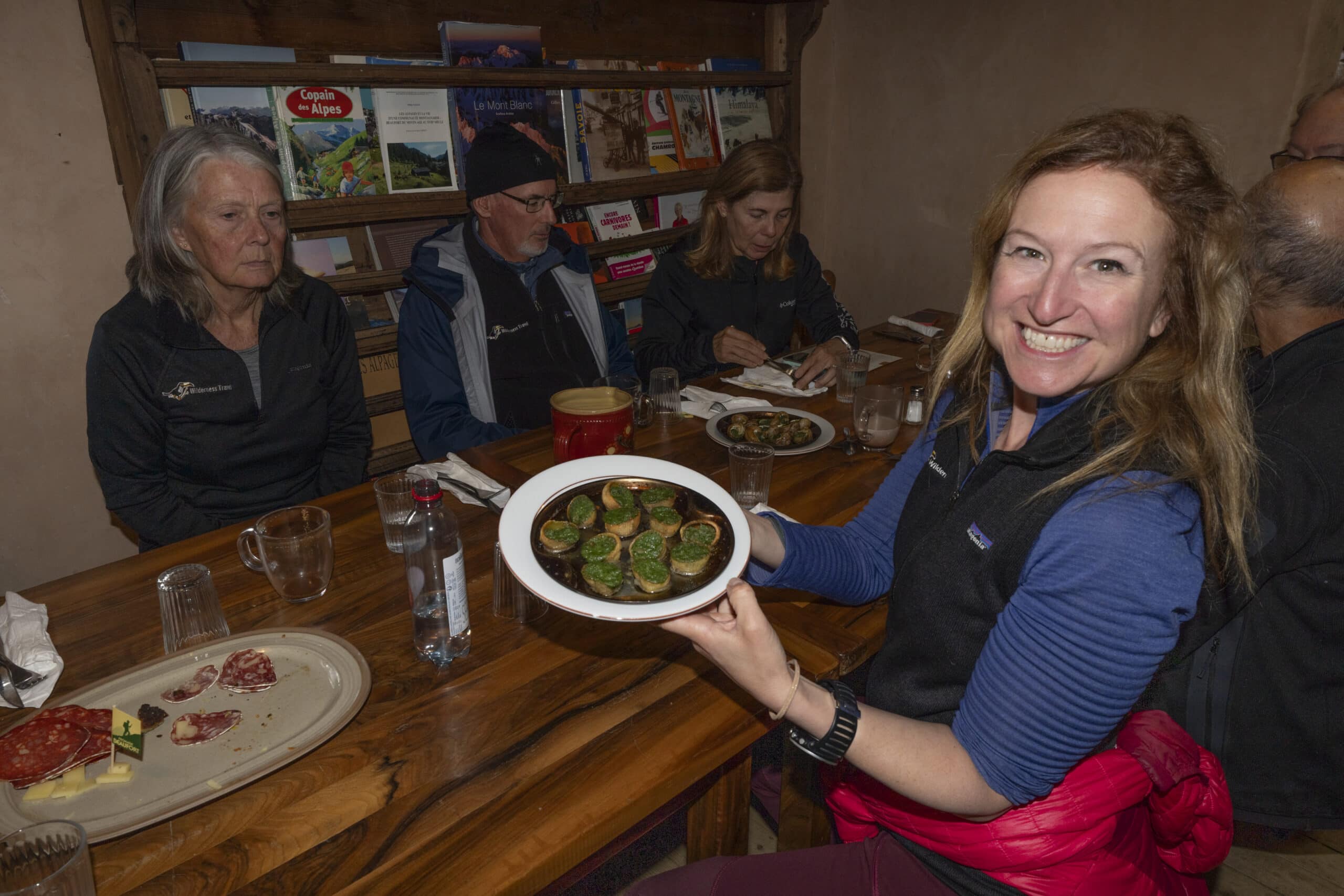 A woman, beaming with joy, holds a plate of food at a table bustling with others. Books adorn the shelf in the background, hinting at tales of adventures like hiking Mont Blanc in the crisp embrace of autumn.