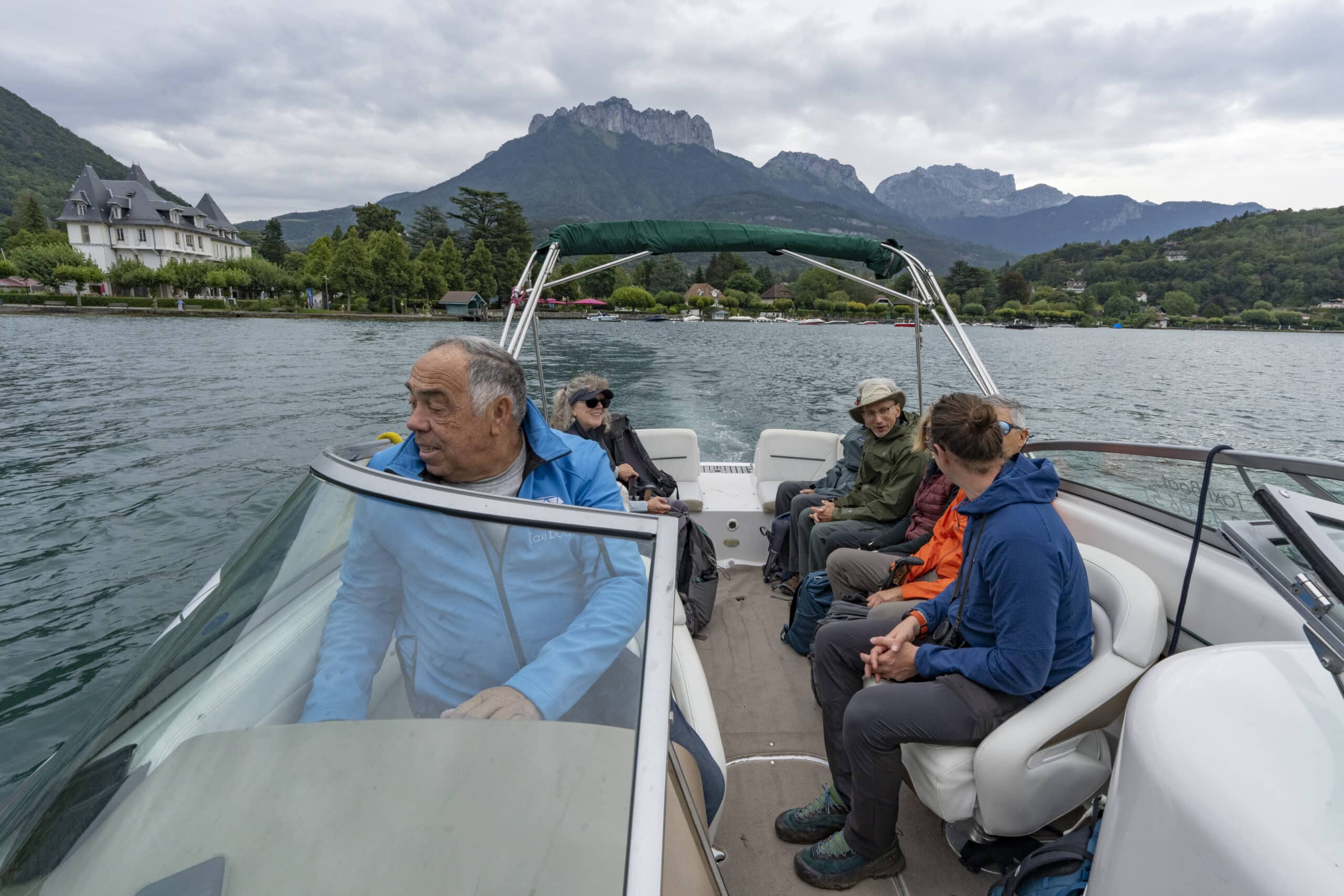 A group of people sit in a motorboat on a lake, surrounded by mountains and a cloudy autumn sky. One person is steering the boat, while others are seated in casual outdoor clothing, discussing their recent hiking adventure near Mont Blanc.