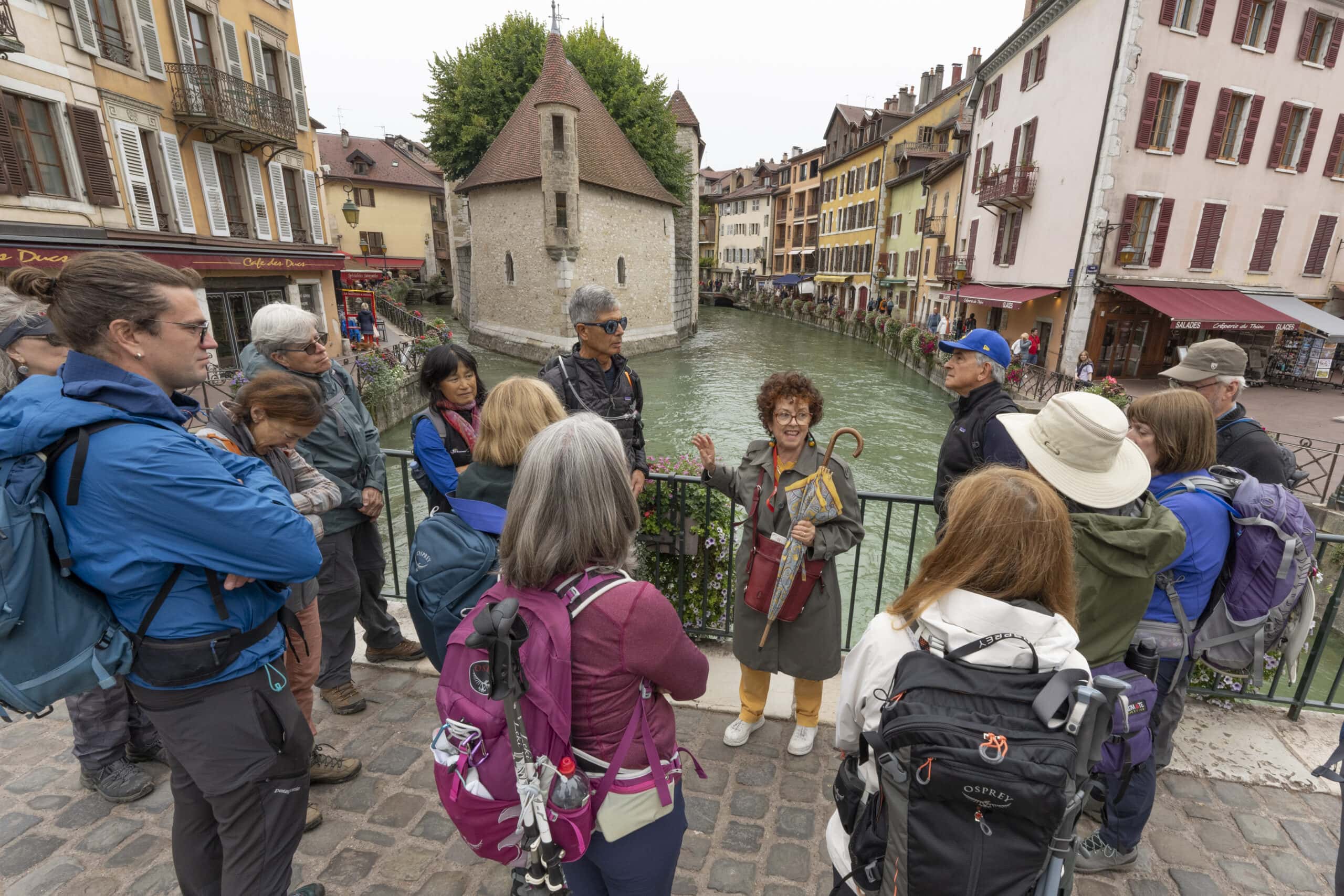 A tour guide stands with a group of tourists near a canal in a European town, surrounded by the vibrant hues of autumn foliage. The colorful buildings and a historic structure provide a picturesque backdrop, creating an unforgettable scene.