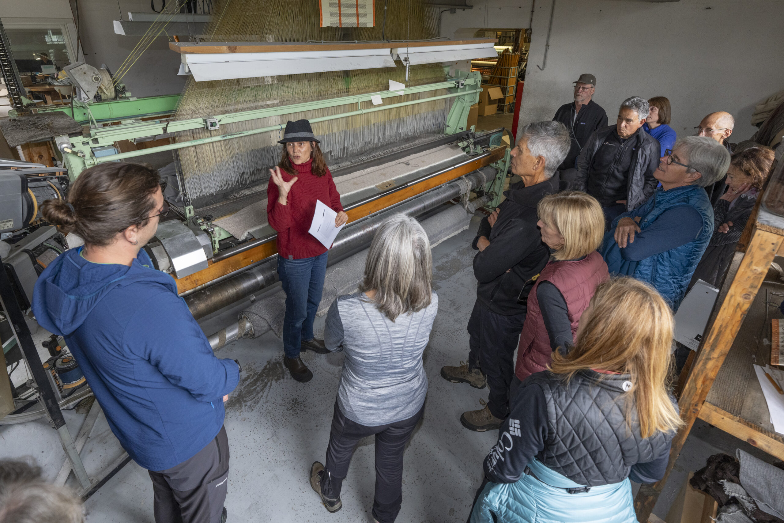 In an autumn-inspired setting, a group stands attentively in a factory as a woman in a red sweater and black hat explains the workings of a large industrial machine, reminiscent of navigating the intricate paths found while hiking near Mont Blanc.