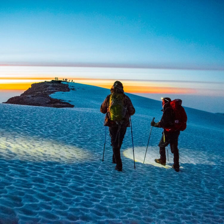 Two hikers with backpacks and trekking poles traverse a snowy mountain ridge at sunrise, capturing one of those important moments as distant people are silhouetted against the colorful 2025 sky.