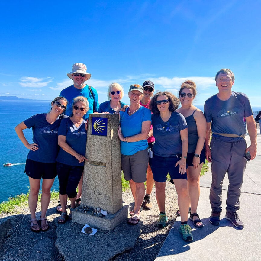 A group of people poses by a stone marker on a coastal path, enjoying the bright blue sky and sea in the background—a snapshot worthy of Moments 2025, capturing the timeless beauty of must-see events along the coastline.