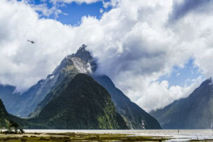 A helicopter flies near tall, cloud-covered mountains beside a body of water with grassy land in the foreground, offering a breathtaking view typical of any Queenstown travel guide.
