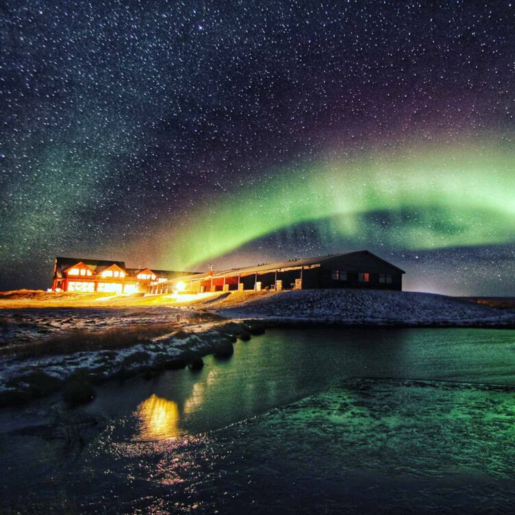 A brightly lit lodge in 2025 sits under a starry sky with the green aurora borealis. Reflections shimmer on a partially frozen pond in the foreground—moments not to miss.