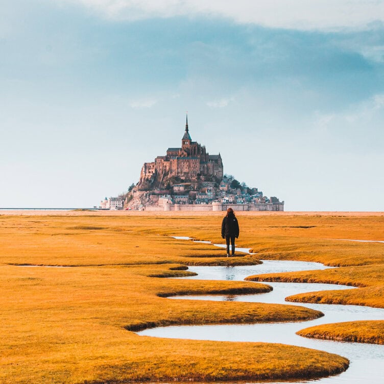 As 2025 unfolds, a person walks on a winding path towards Mont Saint-Michel, surrounded by the not-to-miss beauty of yellow grass beneath a cloudy sky.