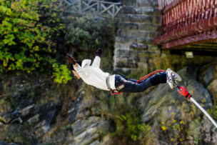 With arms outstretched, a person in a white hoodie bungee jumps off a bridge, surrounded by lush trees and stone walls. If you're looking for top activities, experiencing this thrilling leap in Queenstown is among the unforgettable things to do.