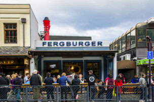 A crowd of people waits outside Fergburger, a popular Queenstown burger joint with a prominent red chimney and adjacent shops—a must-visit for any travel itinerary and one of the unforgettable things to do in town.