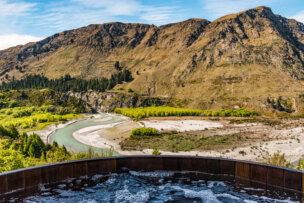 Soak in a hot tub while overlooking Queenstown's attractions—a winding river, vibrant green and yellow trees, and rocky mountains under a clear blue sky.