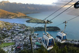 Two gondola cabins ascend a cable over Queenstown's stunning mountainous landscape. Beneath lies a charming town, a shimmering lake, and rolling hills, with majestic peaks in the background—a perfect vantage point for discovering top activities and exciting things to do.