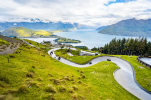 Scenic view of a winding luge track on a grassy hillside overlooking Queenstown's lake, with mountains in the background under a partly cloudy sky—a thrilling activity for those seeking unforgettable things to do.