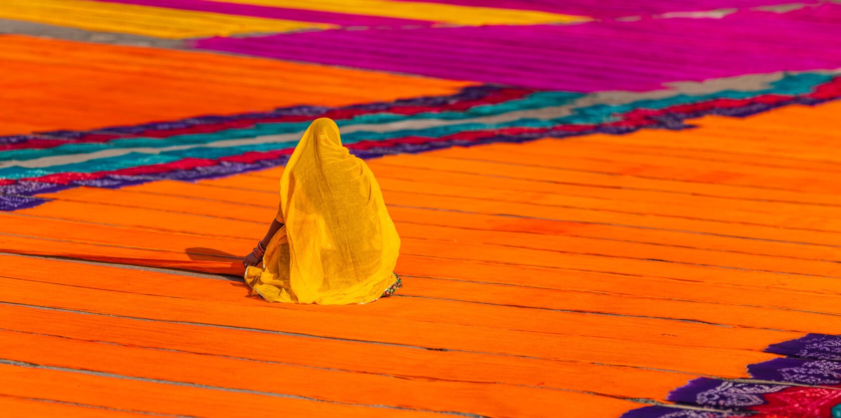 A person draped in a yellow cloth sits among large, vibrant orange and purple fabric pieces spread out on the ground, capturing the vibrant essence that makes an India trip unforgettable.