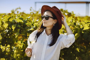 In Queenstown, a woman in a white shirt and red hat enjoys the lush vineyards, sunglasses on and white wine in hand—a perfect snapshot of travel delights and must-see things to do.