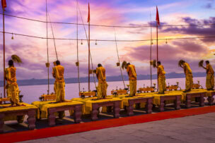 Seven people in traditional attire perform a ceremonial ritual on a platform by the water at sunset in Varanasi, India, with lit lamps and flags in the background.