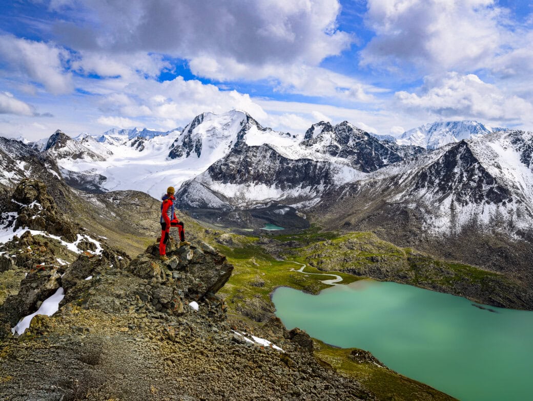 Person in red outdoor gear stands on a rocky cliff overlooking a turquoise lake and snow-capped mountains under a cloudy sky.