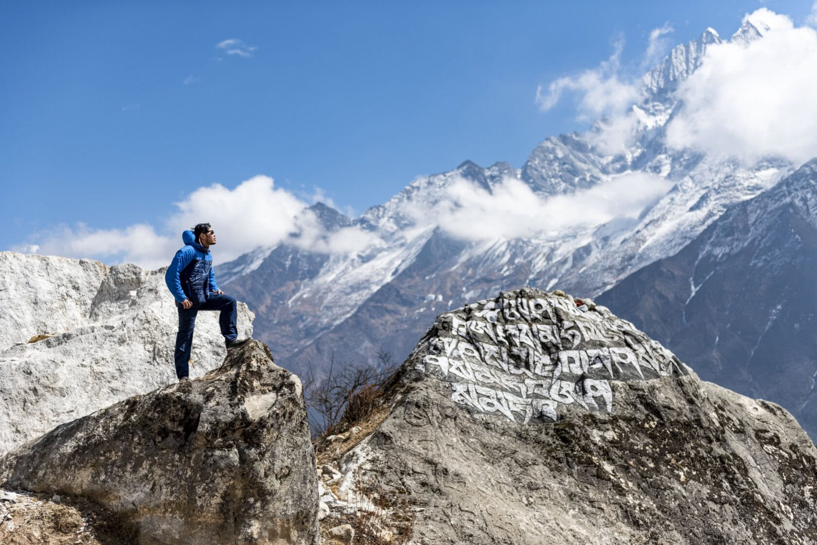 Person in blue hiking gear stands on a rock, looking at snow-capped mountains. Nearby boulder has white inscriptions. Clear blue sky in the background.