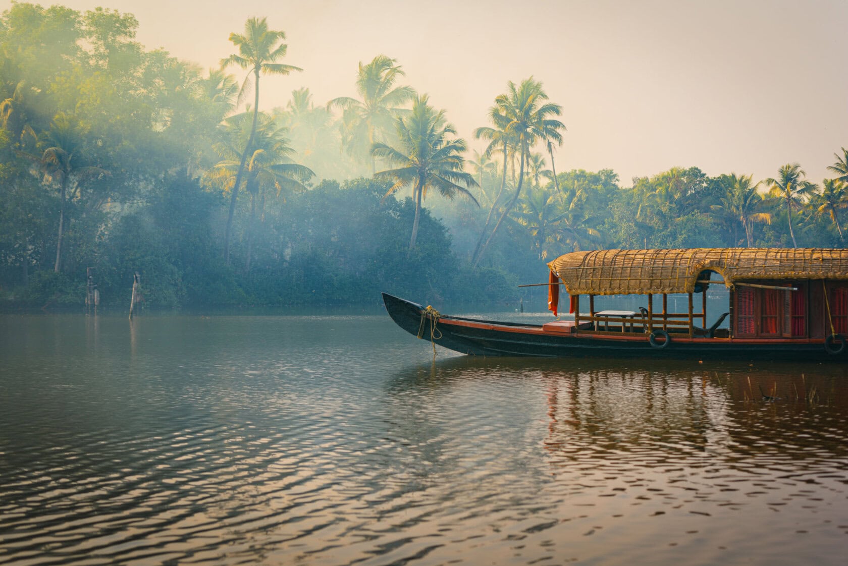 A traditional houseboat floats on a calm river, surrounded by lush tropical vegetation and palm trees under a hazy sky—an enchanting scene that captures the essence of India travel.