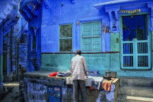 A person stands in front of a vibrant blue building in Rajasthan, India, arranging clothes on a table outside. The building features a small window and a door with bars, offering a colorful glimpse into the everyday life of this culturally rich region.